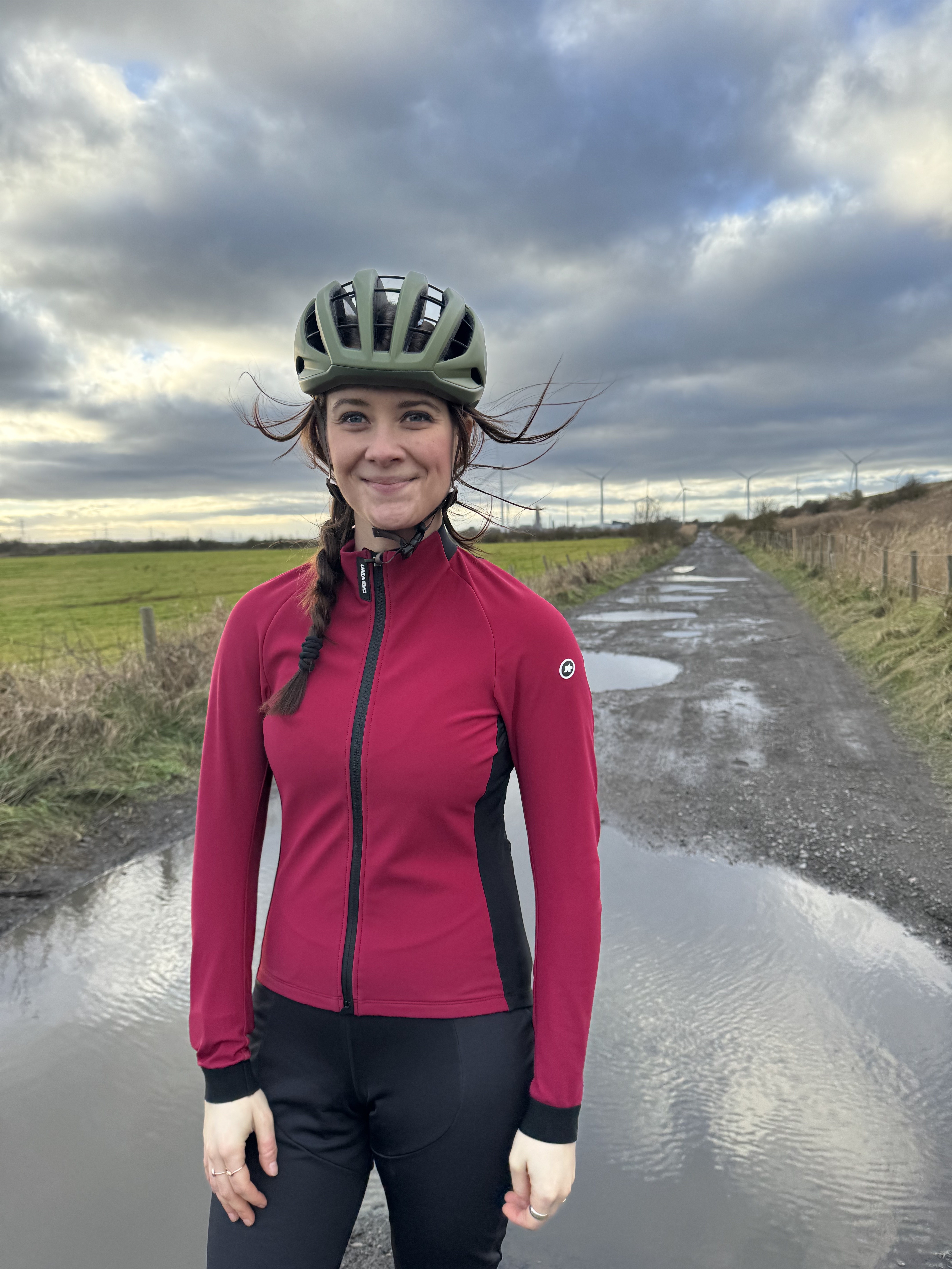 Une femme fait face à la caméra en veste rose et casque vert devant une flaque boueuse.