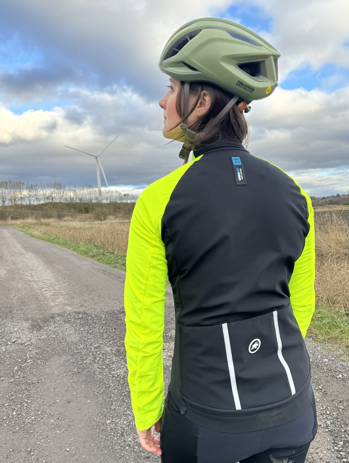 Une femme regardant loin de la caméra dans une veste jaune fluo et un casque vert.  Le dos de la veste est noir avec de grandes poches et des détails réfléchissants.